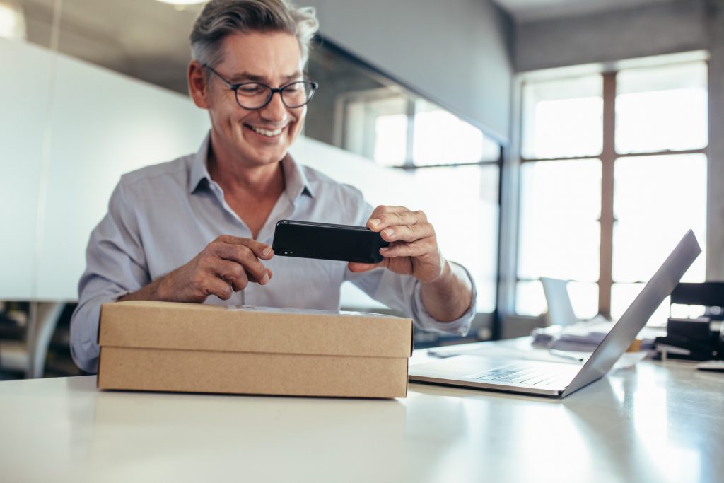 Smiling mid adult man taking scanning a delivery box on his desk. Man taking picture of a package.