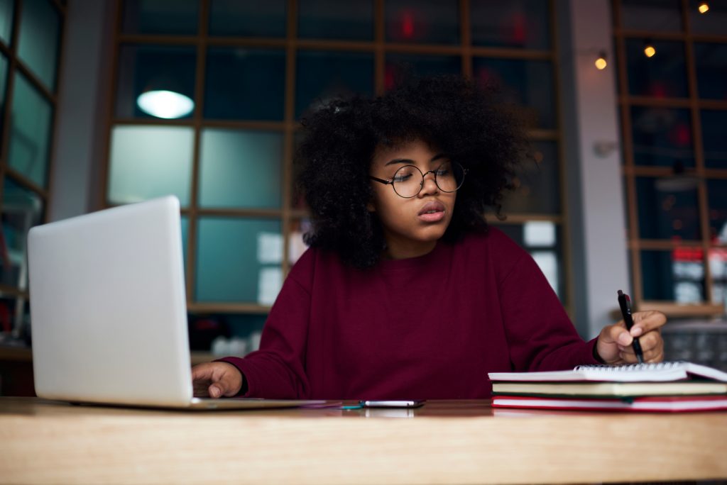 Concentrated young female student in glasses learning in university campus using laptop computer and wireless connection to internet preparing to examination, attractive hipster girl writing essay